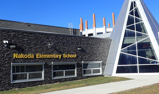 A photograph of the Nakoda Elementary School from the Stoney First Nations Reserve in Alberta, highlighting its modern  steel architectural entrance, which resembles a tipi, against a clear blue sky. Photo by Norman Vaughan, 2019.