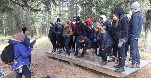 In the woods, a group of adult students stand on a large balance board, with the Fulcrum set fairly close to middle. An older woman, presumably the instructor, and another person stand by. 